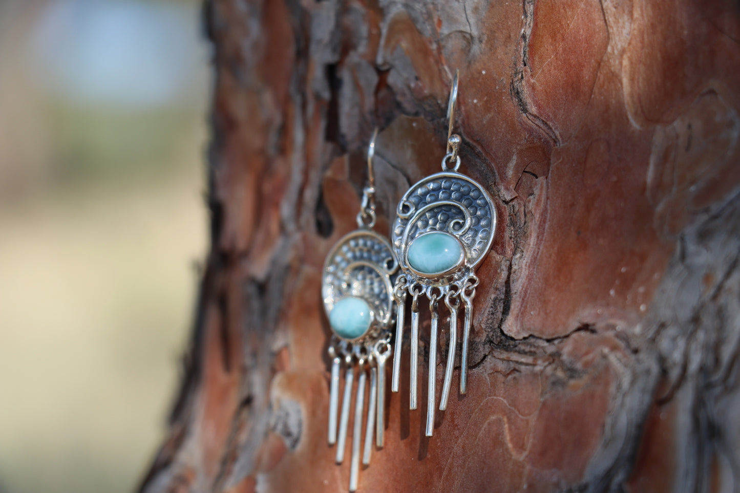 Close-up of handcrafted Larimar earrings in 925 sterling silver, featuring sky-blue oval gemstones and silver fringe design, photographed against tree bark. Natural blue gemstone jewelry.