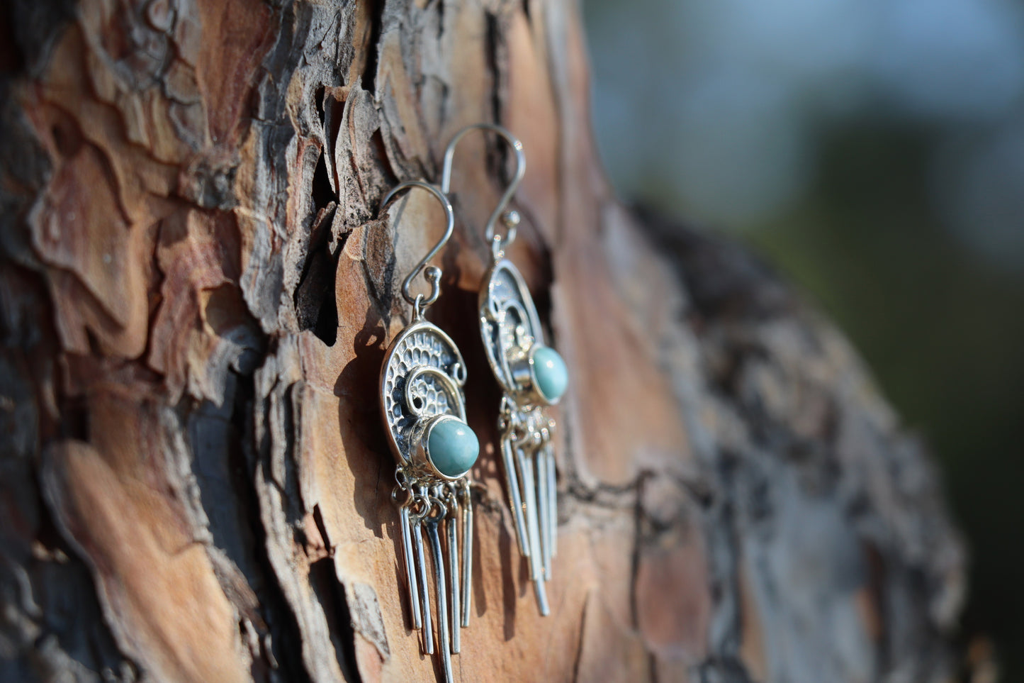 Side view of sterling silver Larimar dangle earrings with soft blue stones and hammered swirl details, hanging on textured tree bark. Artisan-made crystal earrings for calm and clarity.