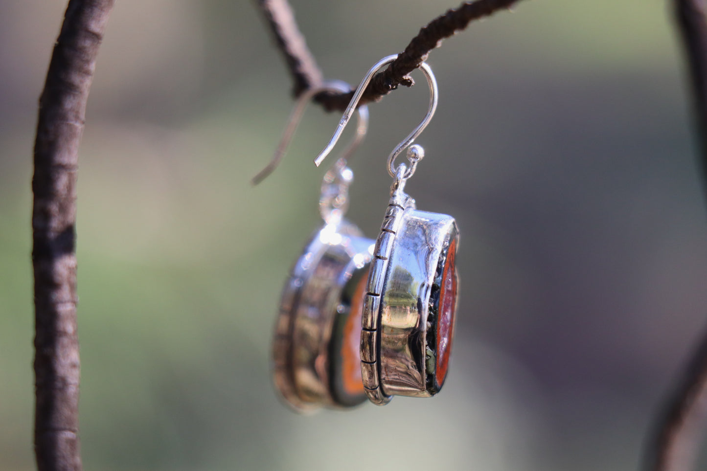 Close-up of a single Tabasco Geode earring handcrafted in 925 sterling silver, showing the vivid red volcanic rim and inner quartz crystal cavity. Hanging from a branch in natural light, highlighting the stone’s Saturn and Pluto energetics for grounding and transformation.