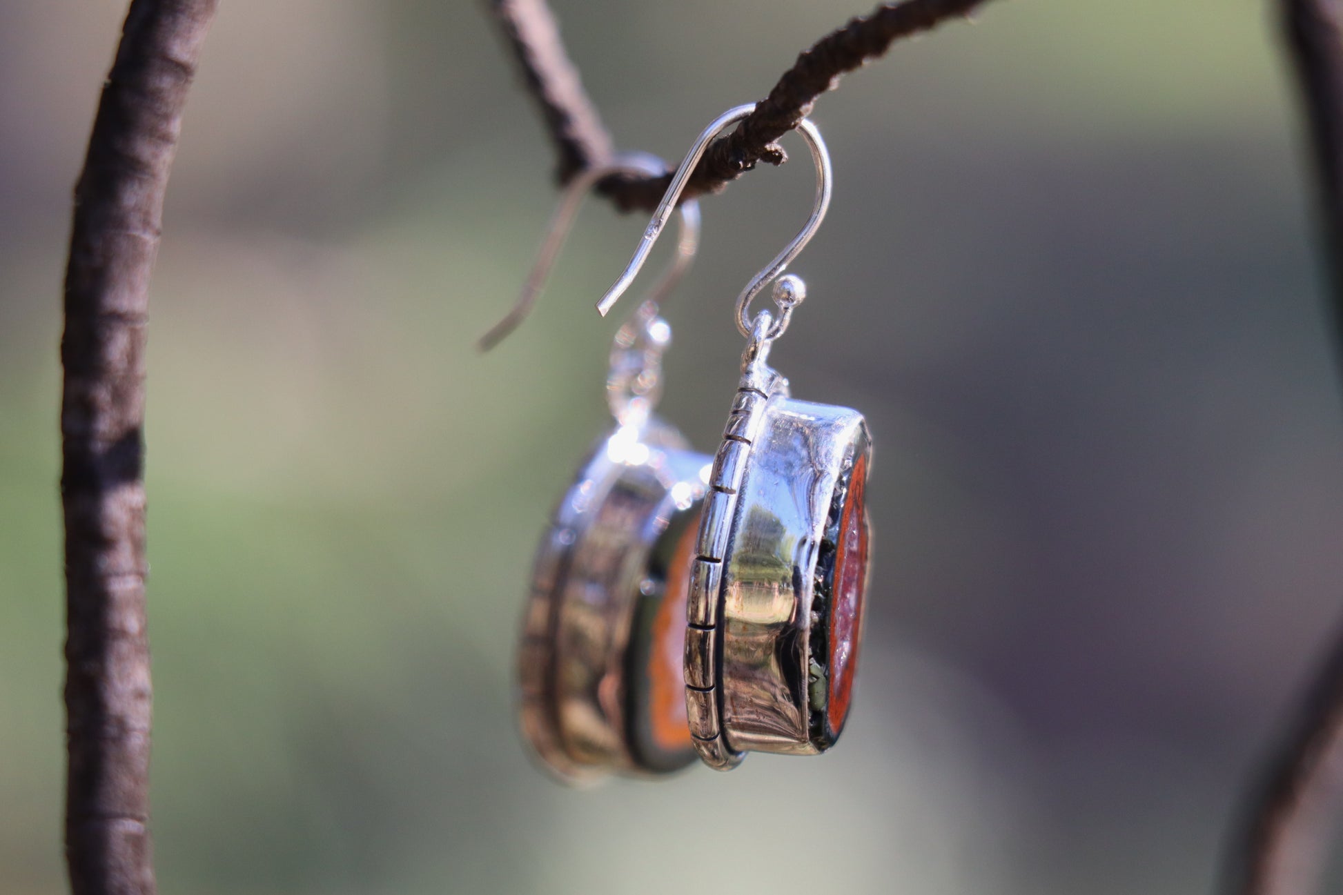 Close-up of a single Tabasco Geode earring handcrafted in 925 sterling silver, showing the vivid red volcanic rim and inner quartz crystal cavity. Hanging from a branch in natural light, highlighting the stone’s Saturn and Pluto energetics for grounding and transformation.